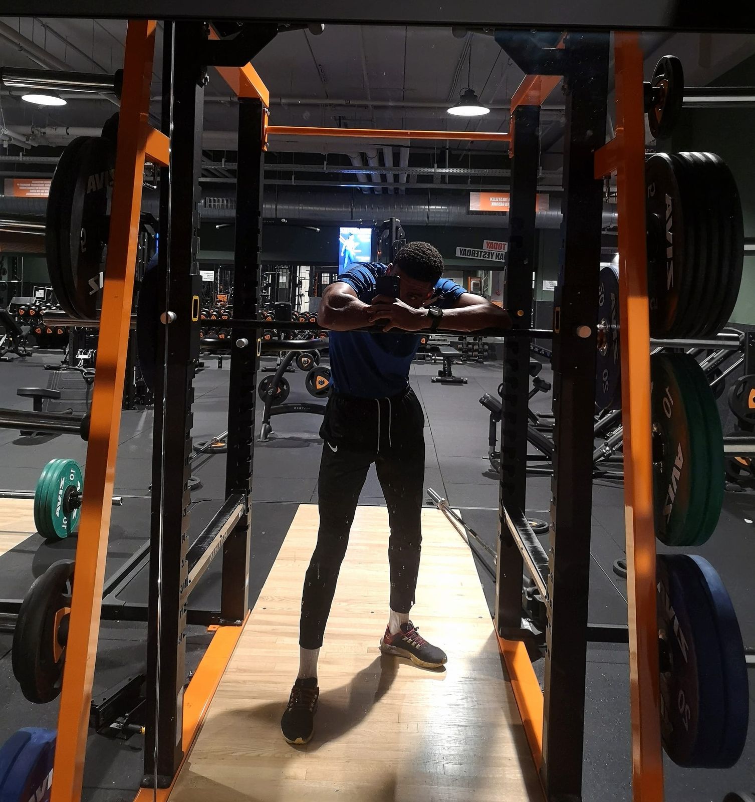 Manu en el gimnasio, dentro de un rack y apoyado en la barra, con camiseta azul y discos alrededor