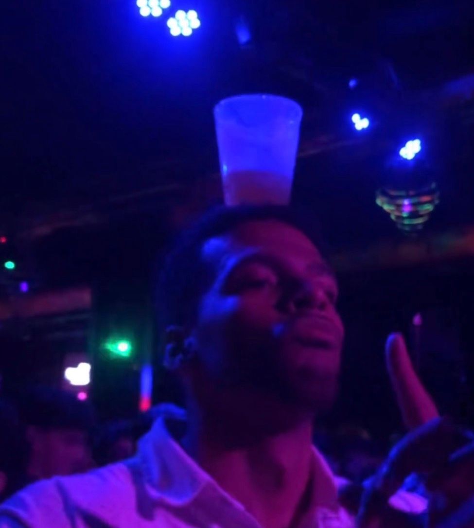Manu de fiesta en una discoteca con luces azules y un vaso en equilibrio sobre la cabeza