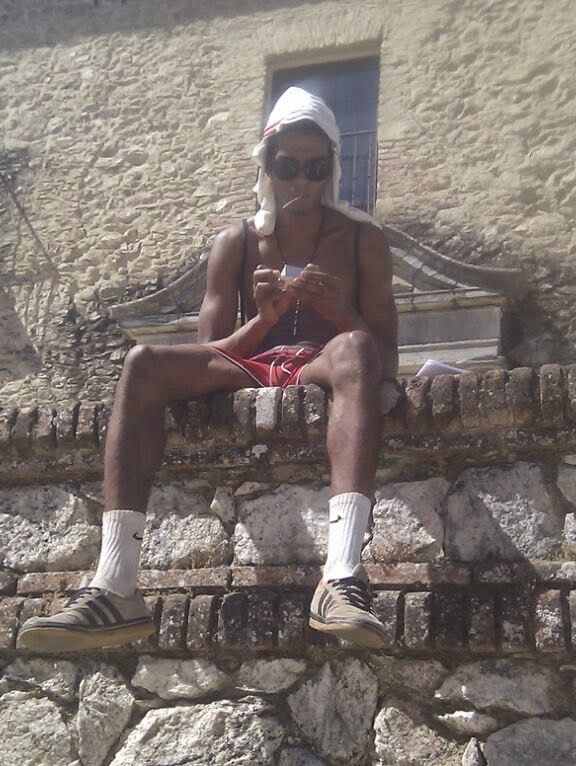 Manu con 63 kg, muy delgado, sentado en un muro de piedra con gorro y gafas de sol